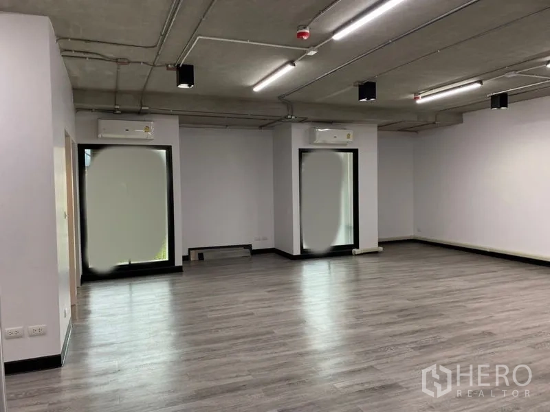 Office Space for rent - Spacious work area with multiple LED lights, exposed ceiling, and two tall windows fitted with AC units.