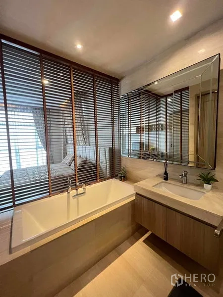 Condominium for rent or sale in Watthana, Bangkok - Bathroom with soaking tub next to louvered glass partition and a wide vanity mirror.