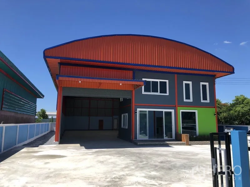 Warehouse for rent or sale in Sai Noi, Nonthaburi - Modern two-tone warehouse exterior with orange roof, wide roller door, glass-fronted office and large concrete yard.
