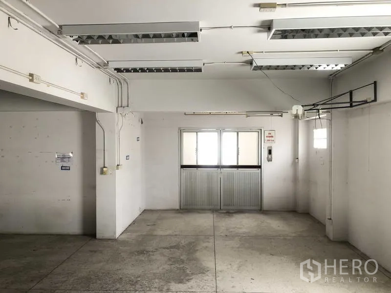 Factory for rent in Prawet, Bangkok - Industrial room with fluorescent lighting, exposed power lines and a sliding door.