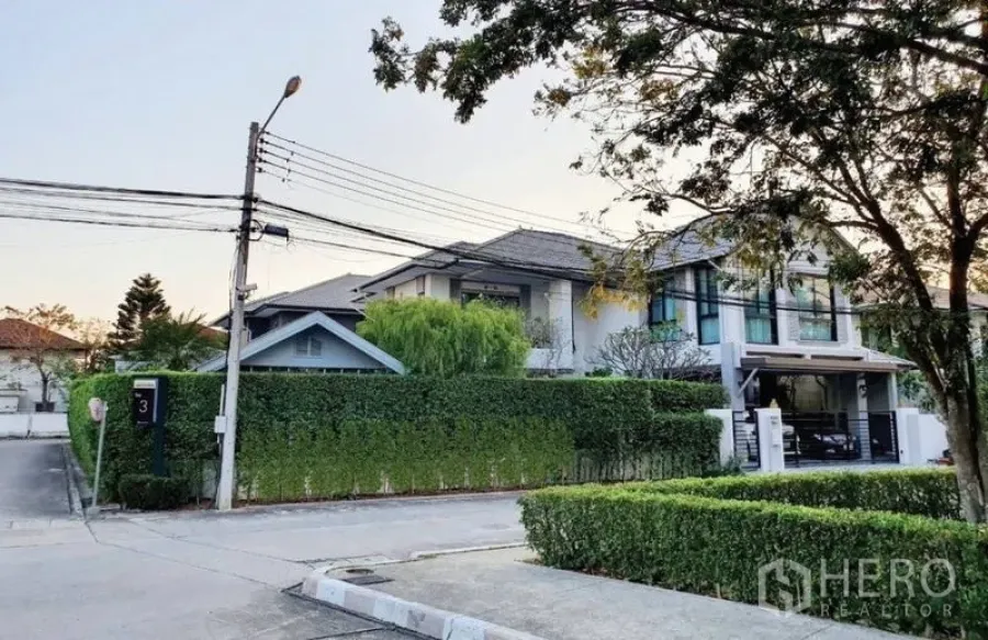 Detached House for sale in Lam Luk Ka, Pathum Thani - Corner-lot two-storey detached house with tall green hedge, carport and tree-lined street.