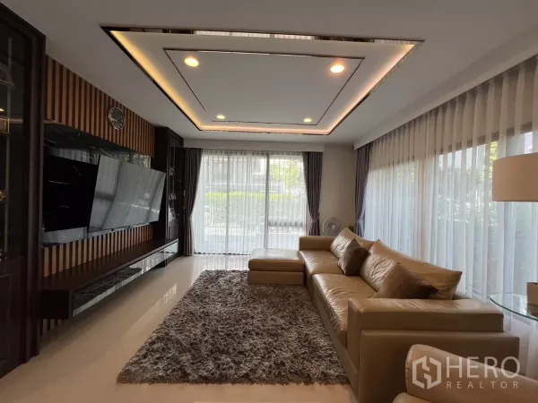 Detached House for rent or sale in Bang Kapi, Bangkok - Living room with L-shaped leather sofa, large wall-mounted TV and cove lighting.
