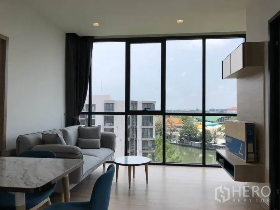 Condominium for rent in Watthana, Bangkok - Bright living room with floor‑to‑ceiling windows, sofa and dining area overlooking a canal and city skyline.