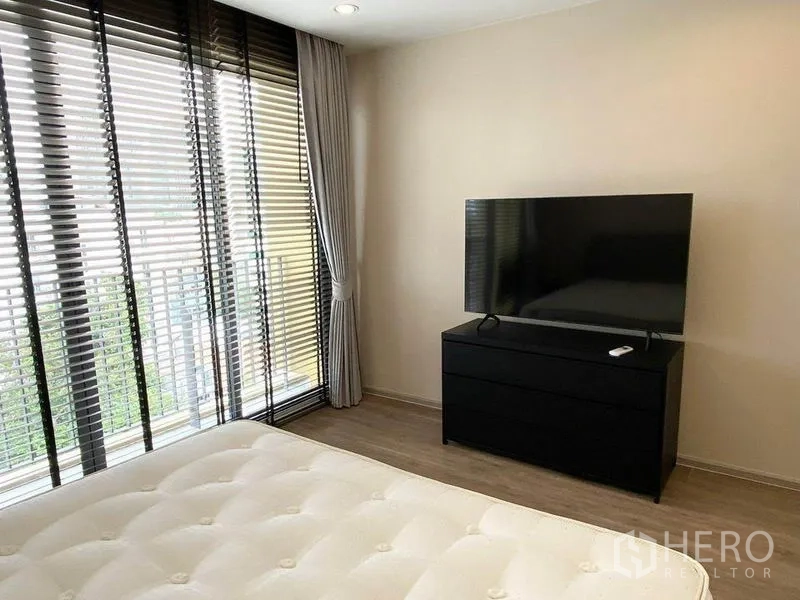 Condominium for rent in Pathum Wan, Bangkok - Bedroom corner with large windows, blackout curtains and TV on a black chest of drawers.