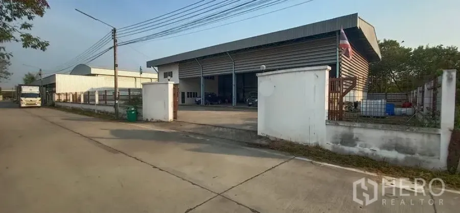 Warehouse for rent in Khlong Luang, Pathum Thani - Gated street-front entrance to a metal-clad warehouse with a wide driveway and perimeter wall in Khlong Luang.