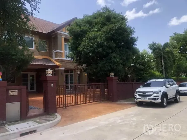 Detached House for rent or sale in Pak Kret, Nonthaburi - Gated driveway and front fence on a quiet estate road with mature trees.