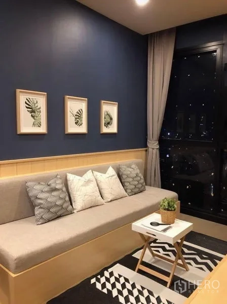 Condominium for rent in Watthana, Bangkok - Cozy sofa nook with a dark blue feature wall, framed botanical art, rug, and night city view through the window.