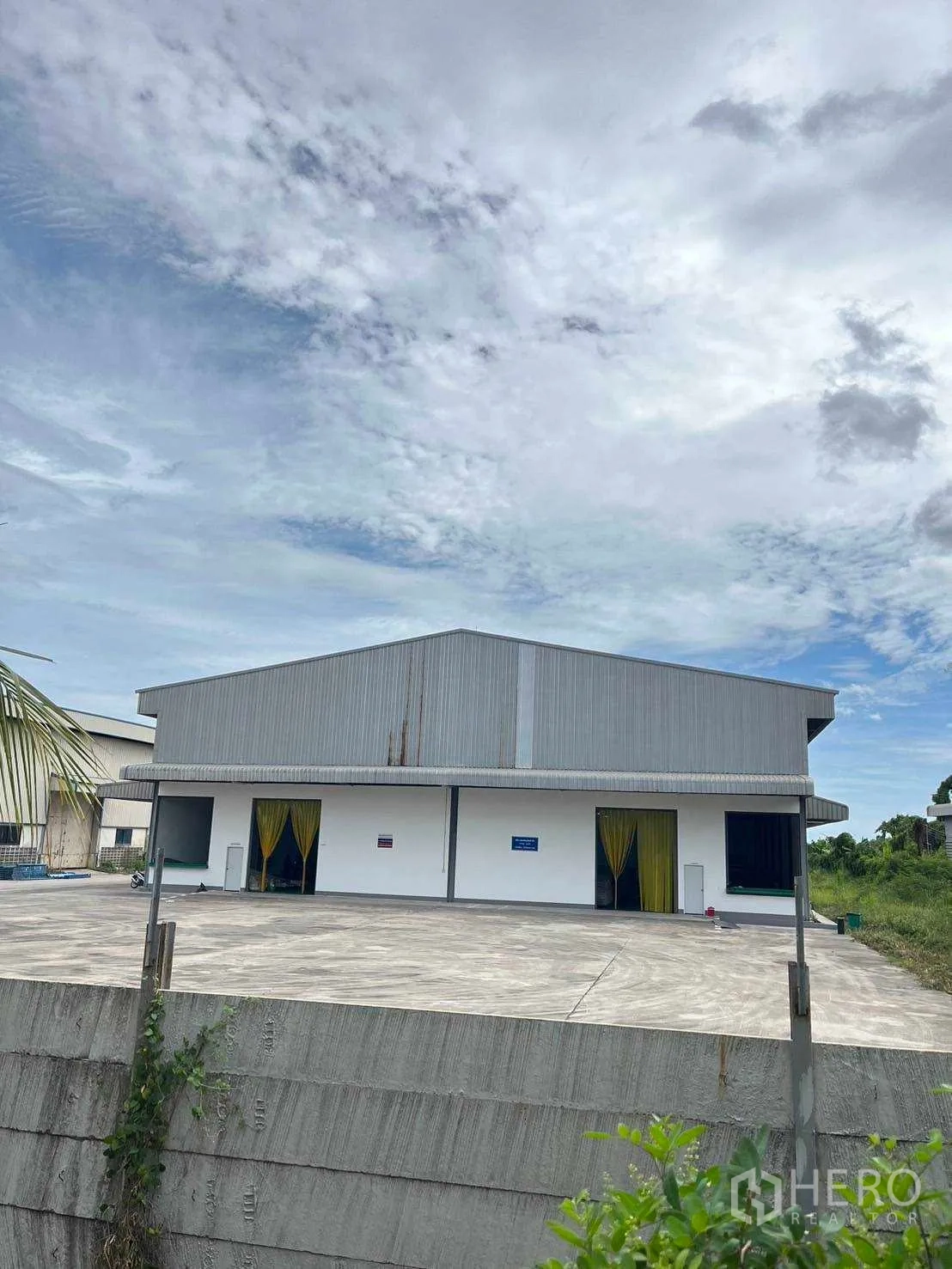 Factory for sale in Nikhom Phatthana, Rayong - Warehouse facade with covered loading area and elevated concrete yard behind a boundary wall.