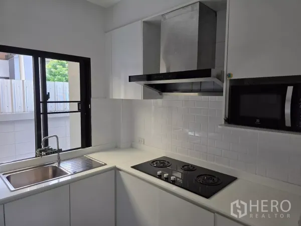 Detached House for rent in Prawet, Bangkok - Clean white L-shaped kitchen with gas hob, range hood, window sink and microwave.