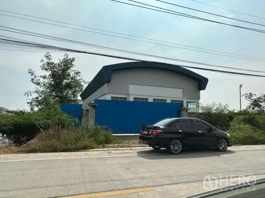 Factory for rent in Pak Kret, Nonthaburi - Street-facing view of a modern factory with blue perimeter wall and arched metal roof along a concrete road.