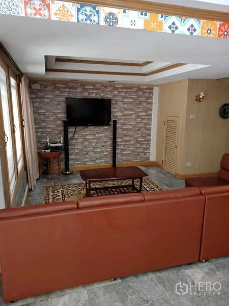 Townhouse for rent in Khlong Toei, Bangkok - Cozy living room with leather sofa, TV on a brick feature wall, and recessed ceiling in an Ekkamai townhouse.