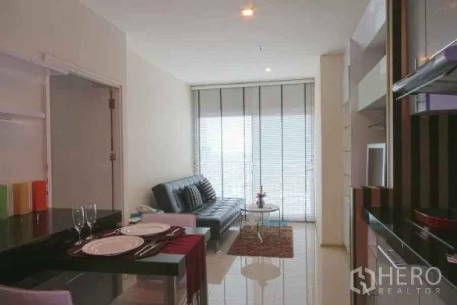 Condominium for rent in Khlong Toei, Bangkok - Bright living and dining area with breakfast bar, leather sofa, and floor‑to‑ceiling windows leading to a balcony.