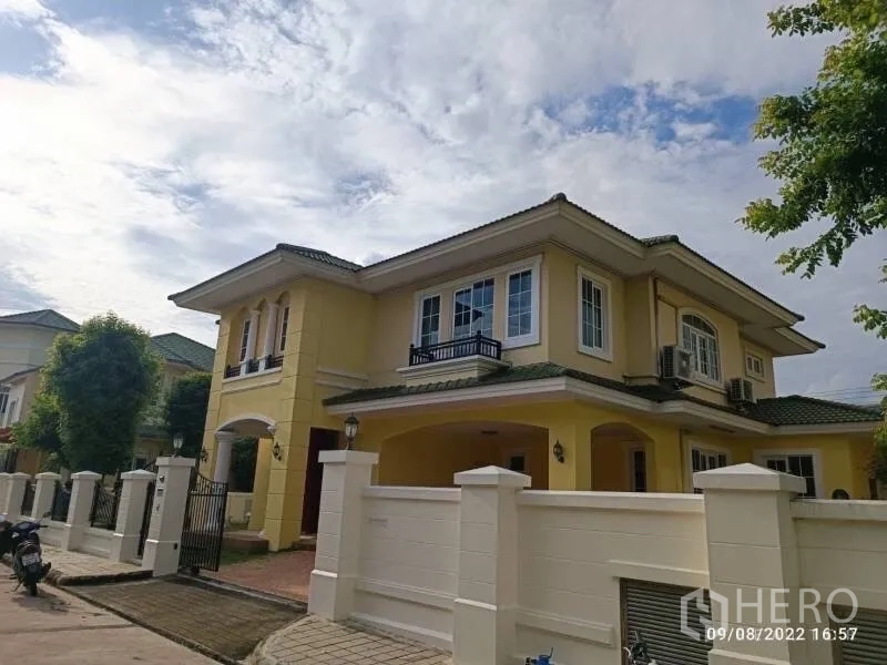 Detached House for sale in Prawet, Bangkok - Front exterior of a two-storey yellow detached house with white fence and carport in Prawet, Bangkok under blue sky.