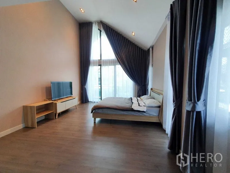 Detached House for rent in Khlong Luang, Pathum Thani - Master bedroom with high ceiling, TV stand and wide glass doors framed by drapes.