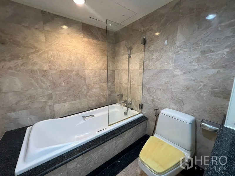 Condominium for rent or sale in Khlong Toei, Bangkok - Marble-tiled bathroom featuring a bathtub with handheld shower and glass screen.