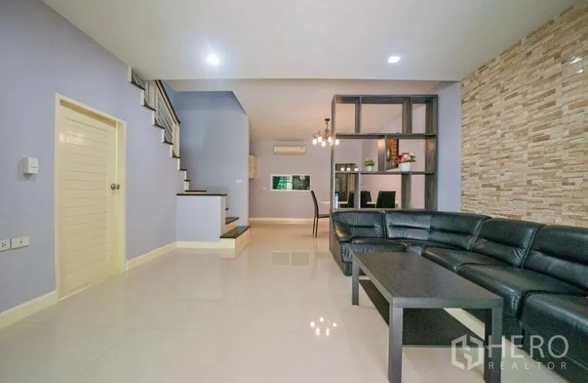 Townhouse for rent in Suan Luang, Bangkok - Open-plan living area with tiled floors, staircase, stone feature wall and a black leather sectional sofa.