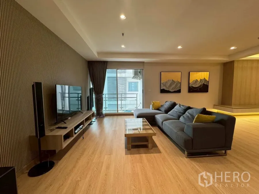 Condominium for rent or sale in Khlong Toei, Bangkok - Spacious living room with TV wall, grey sectional sofa and balcony window.