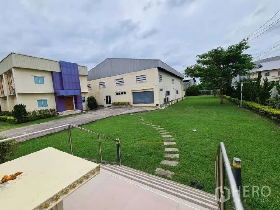 Warehouse for sale in Bang Phli, Samut Prakan - View across the warehouse compound showing driveway, roller-shutter loading bay, and a large grassy yard.