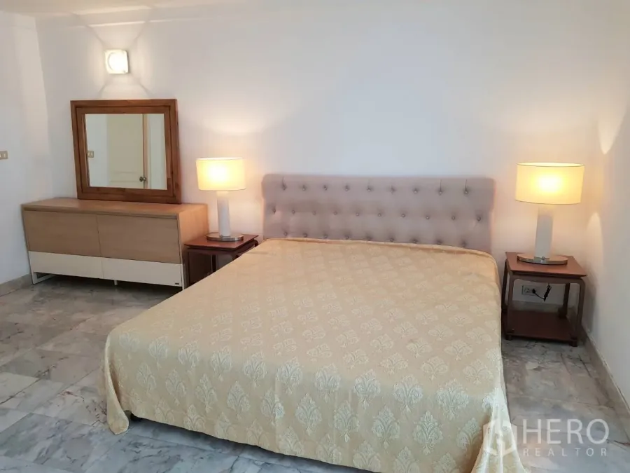 Condominium for rent in Khlong Toei, Bangkok - Primary bedroom with upholstered headboard, bedside lamps and dresser mirror on marble flooring.