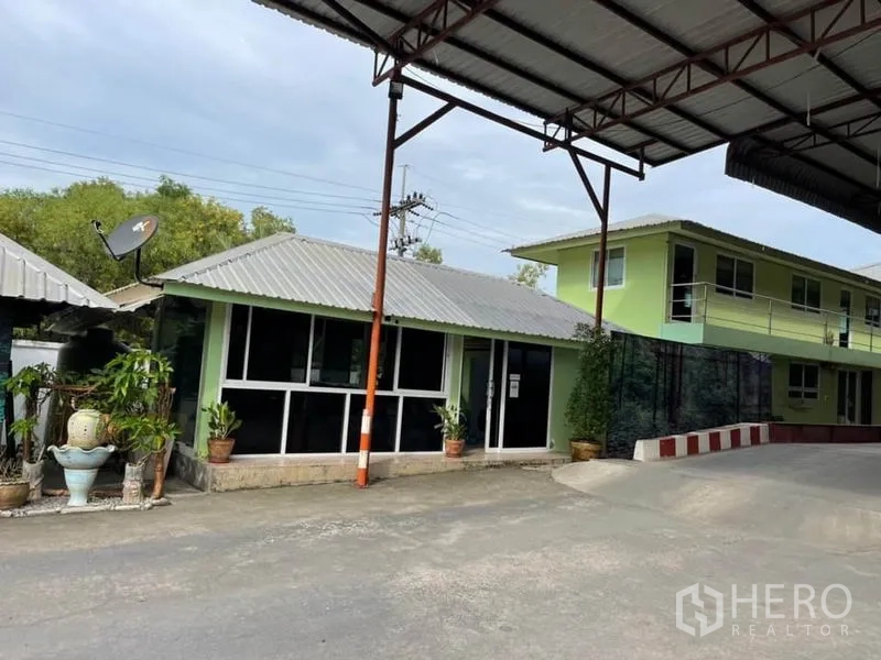 Warehouse for sale in Lam Luk Ka, Pathum Thani - On-site single- and two-story office buildings beneath a metal canopy with large windows and potted plants.
