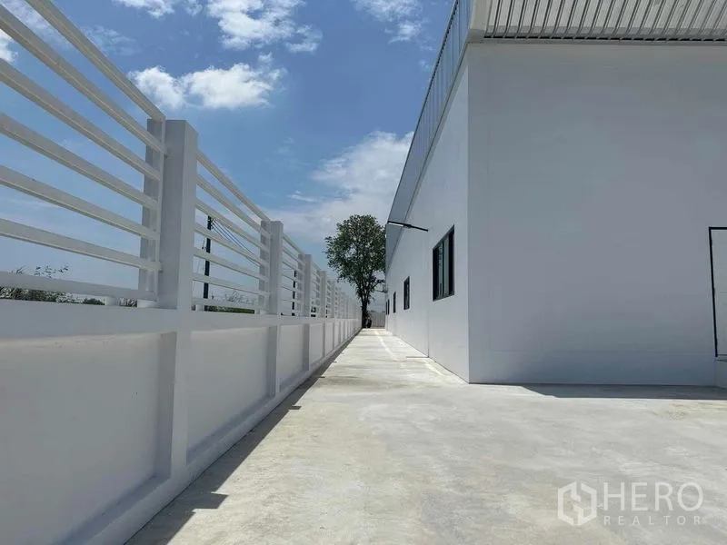 Warehouse for sale in Khlong Luang, Pathum Thani - Side walkway with white perimeter fence running along the warehouse under blue sky.