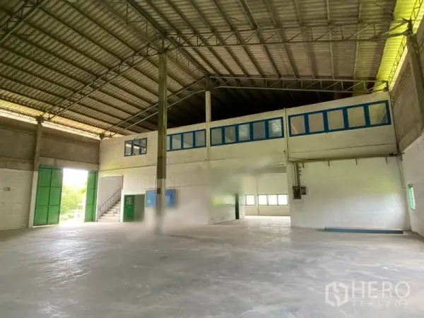 Warehouse for rent in Khlong Sam Wa, Bangkok - Warehouse hall showing blue-framed office windows, large green steel access doors, and a wide clear-span floor.