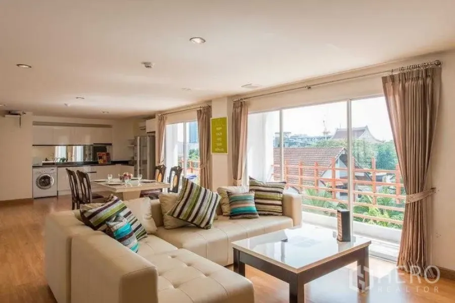 Condominium for rent in Khlong Toei, Bangkok - Open living room with L-shaped sofa, coffee table and balcony windows over low-rise rooftops.