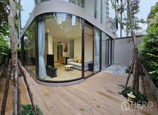 Condominium for rent or sale - Ground-floor condo with curved floor-to-ceiling glass and a private wooden terrace bordered by greenery.