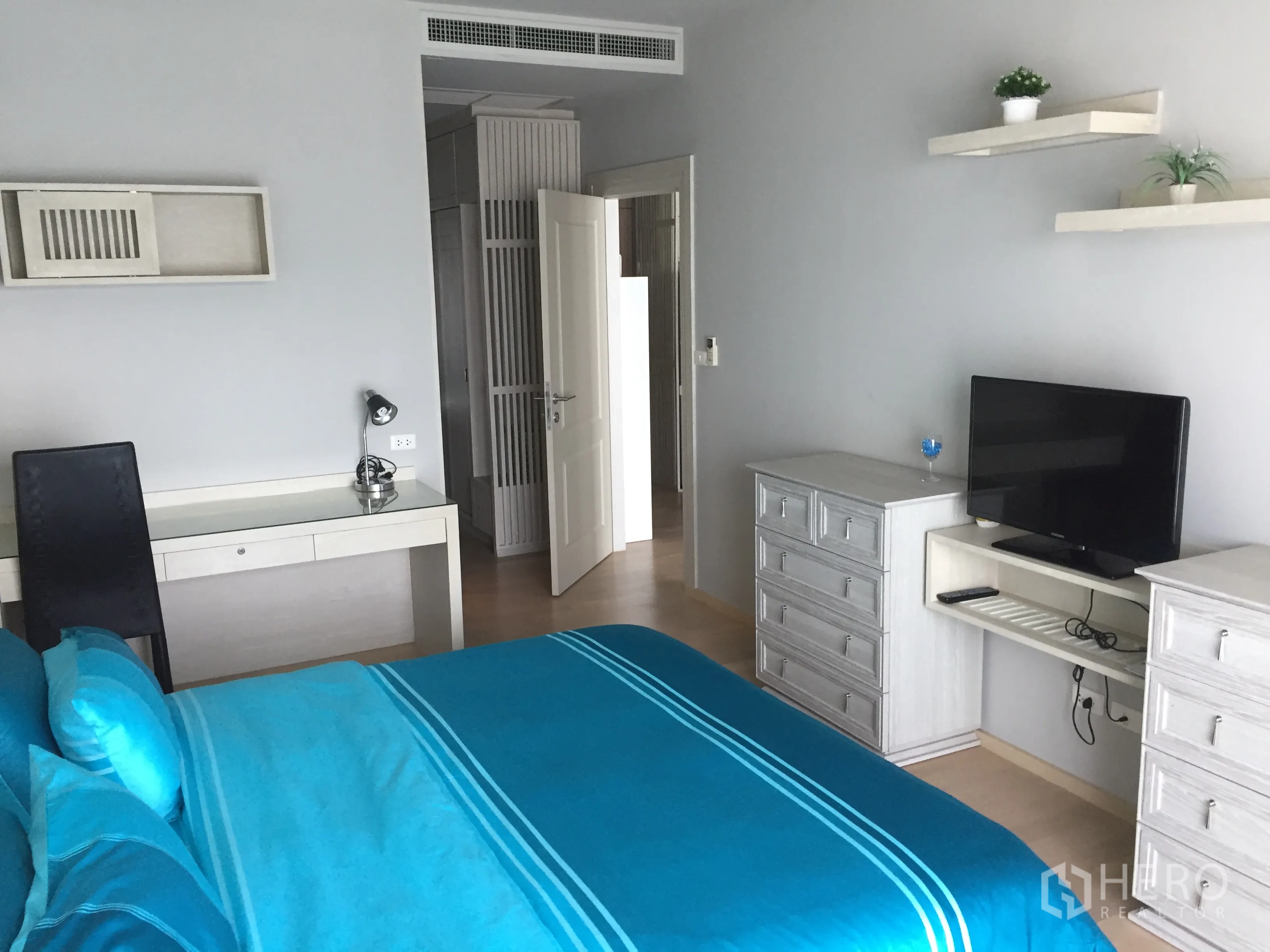 Condominium for rent in Bangkok - Cozy bedroom with blue bedding, work desk, drawers, wall shelves and a flat-screen TV.