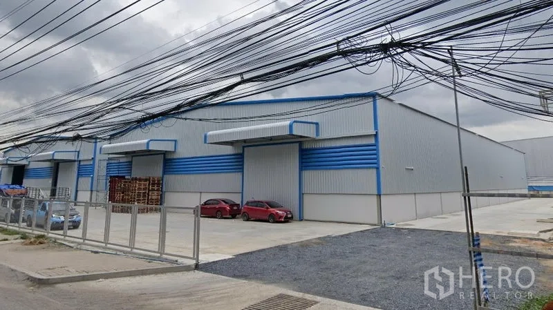 Warehouse for rent in Bang Phli, Samut Prakan - Gated warehouse complex with multiple bays, parking spaces and overhead utility lines along the road.