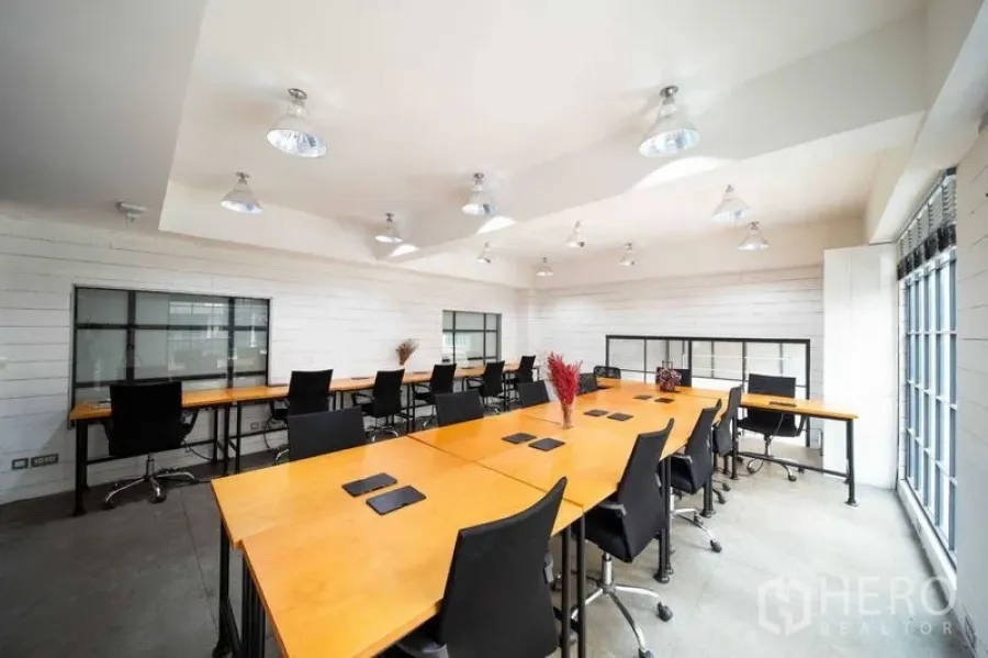 Office Space for rent - Bright open-plan office with long wooden desks, black chairs, white walls, and large windows in the Thonglor area.