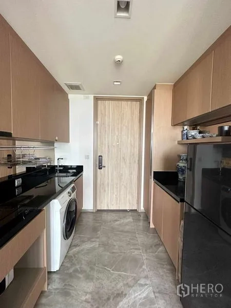 Condominium for sale in Watthana, Bangkok - Galley kitchen fitted with black stone countertops, built‑in cabinets, refrigerator, and washing machine.