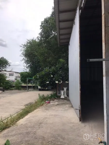 货栈 出租在Bang Kapi, Bangkok - 仓库金属外墙侧立面，宽幅推拉门，车道出入方便，周边有绿植。