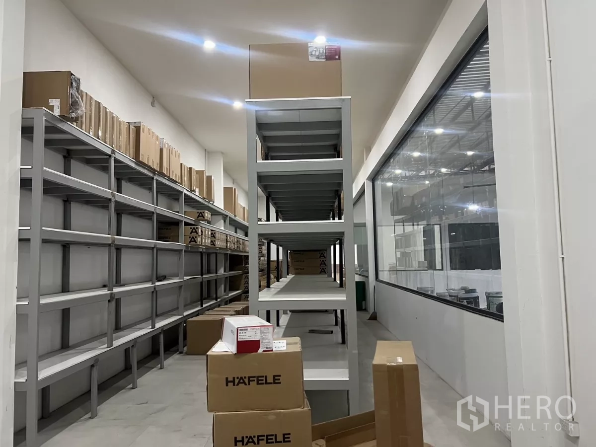 Warehouse for rent or sale in Lat Krabang, Bangkok - Aisle with metal shelving and boxes next to a large internal glass window.
