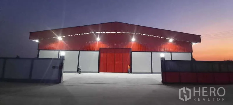 Warehouse for rent or sale in Khlong Luang, Pathum Thani - Front elevation at dusk showing wide red sliding doors, bright floodlights and a spacious driveway.
