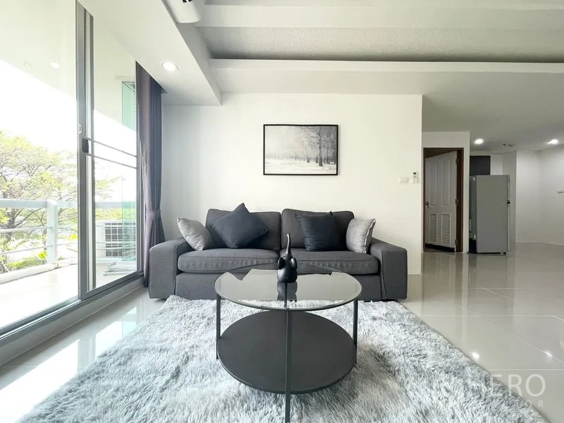 Condominium for rent in Khlong Toei, Bangkok - Living room with grey sofa on a plush rug beside the balcony.