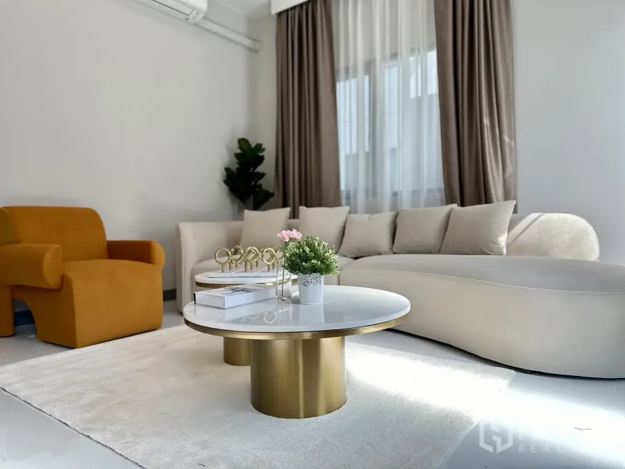 บ้าน ให้เช่าหรือขาย - ห้องนั่งเล่นสว่างพร้อมโซฟาโค้งสีครีม เก้าอี้สีมัสตาร์ด โต๊ะกลางหินอ่อนขอบทอง และผ้าม่านโปร่ง