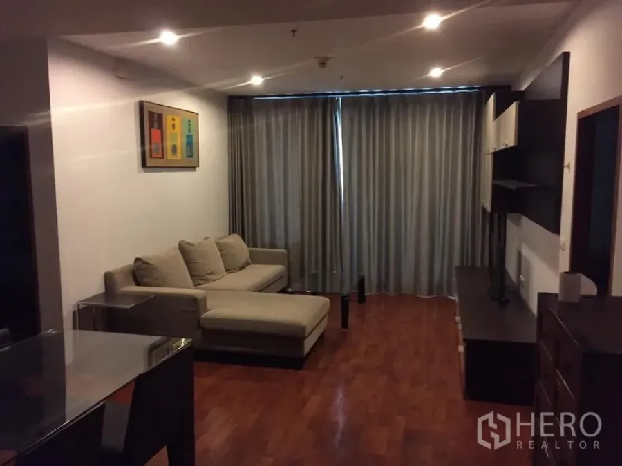 Condominium for rent or sale in Khlong Toei, Bangkok - Cozy living room with L-shaped sofa, wood flooring, media wall and full-length blackout curtains.