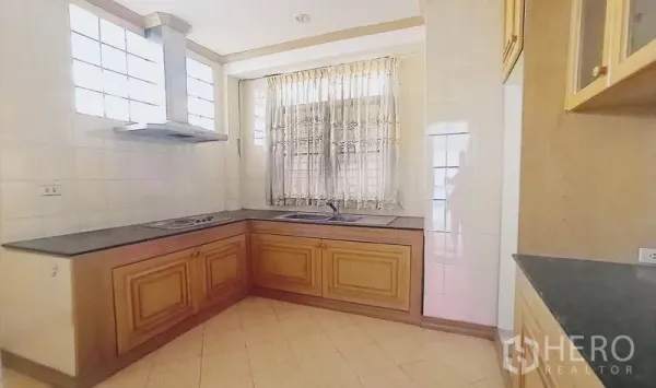 Detached House for sale in Bang Kapi, Bangkok - L-shaped kitchen with wood cabinetry, cooktop, range hood and curtained window.
