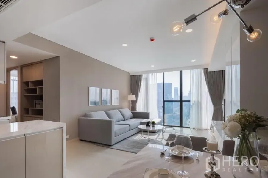 Condominium for rent in Khlong Toei, Bangkok - Bright open-plan living and dining area with a gray sofa, marble island, and floor-to-ceiling windows with city view.