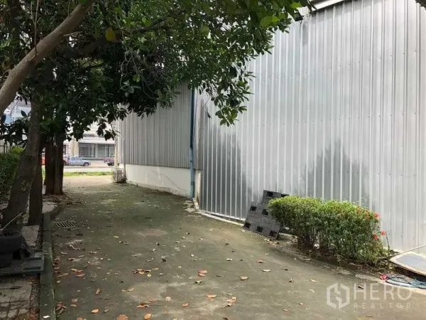 Warehouse for rent in Pak Kret, Nonthaburi - Shaded service lane between corrugated metal buildings with greenery and concrete walkway.