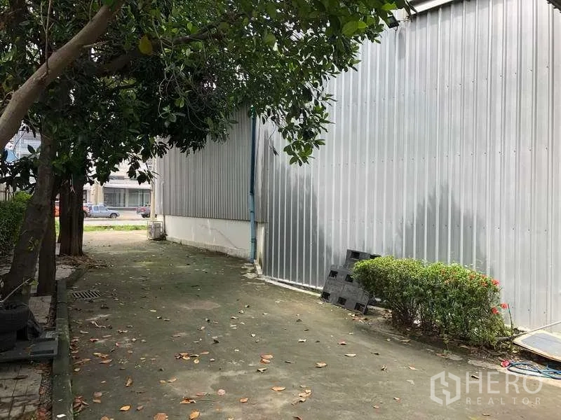 Warehouse for rent in Pak Kret, Nonthaburi - Shaded service lane between corrugated metal buildings with greenery and concrete walkway.