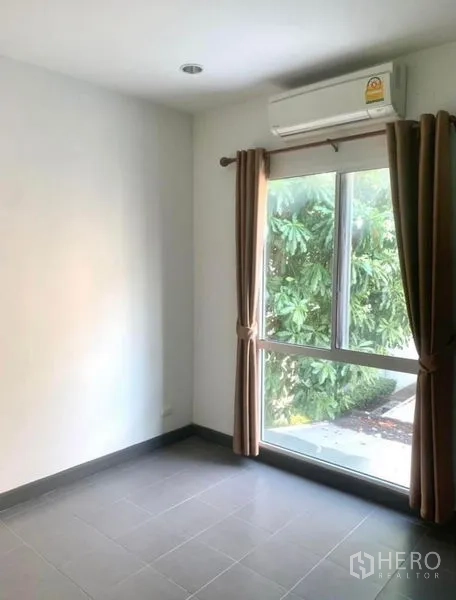Townhouse for rent in Suan Luang, Bangkok - Ground-floor room with large window, brown curtains and split air conditioner facing greenery.