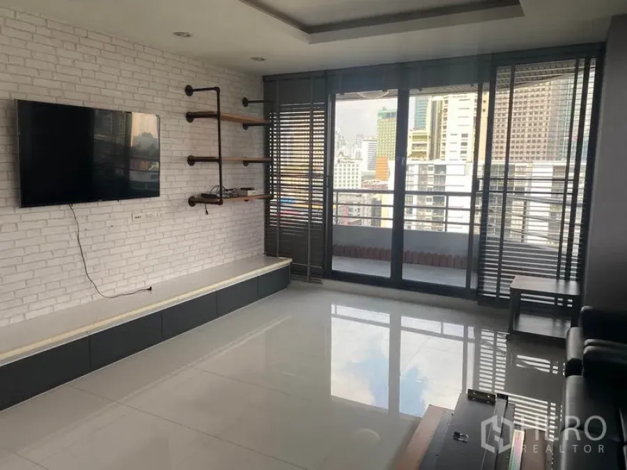 Condominium for rent in Khlong Toei, Bangkok - Living room with balcony access, white brick feature wall, mounted TV, and skyline view.