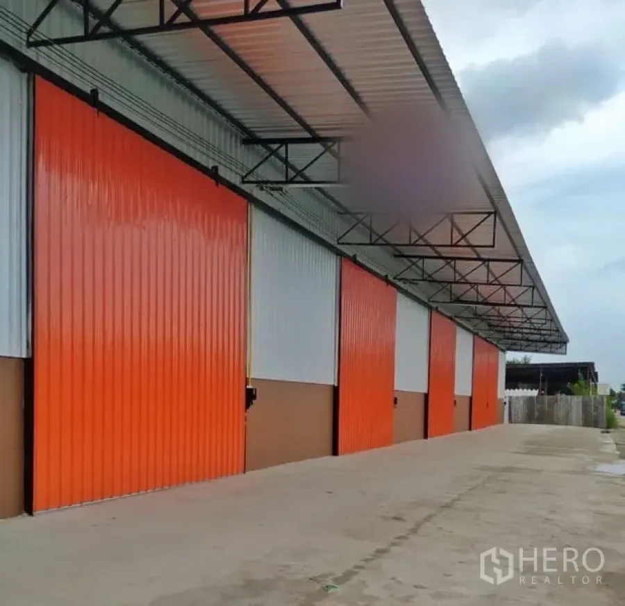 Warehouse for rent in Khlong Luang, Pathum Thani - Row of modern warehouses with orange sliding doors, wide concrete driveway, and a large covered loading canopy.