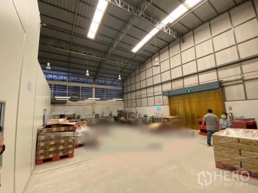 Warehouse for rent in Bueng Kum, Bangkok - High-ceiling warehouse interior with steel trusses, louvered windows, pallets, and a yellow PVC strip door.