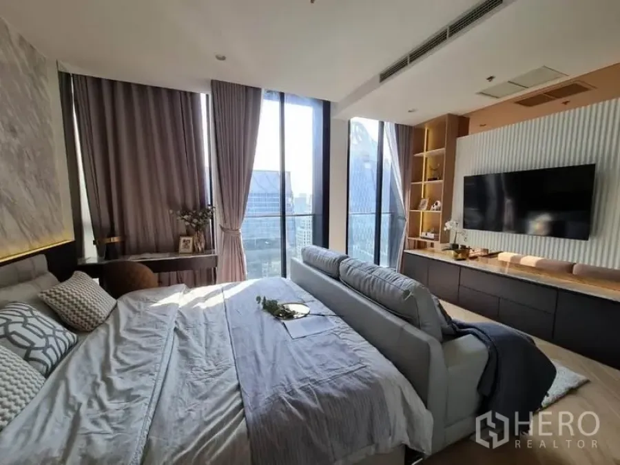 Condominium for sale in Pathum Wan, Bangkok - Sunlit bedroom and lounge with floor-to-ceiling windows, skyline view and built-in TV wall.