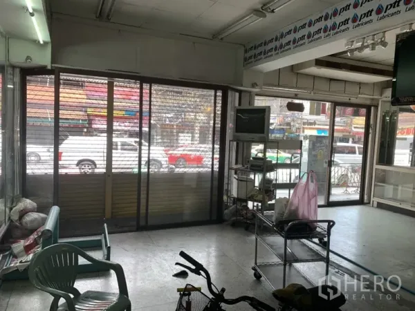 Shophouse for rent in Khlong Toei, Bangkok - Ground-floor retail space with wide glass storefront, metal security gate and fluorescent lighting facing the road.
