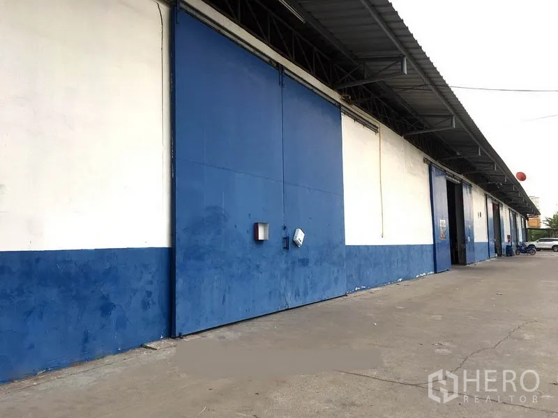 Warehouse for rent in Bang Phli, Samut Prakan - Blue-and-white warehouse exterior with large sliding steel doors and covered loading bay along a wide concrete driveway in Bang Phli.