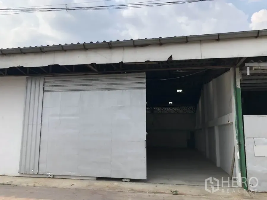 Warehouse for rent or sale in Khlong Luang, Pathum Thani - Front exterior of single-storey warehouse with wide metal sliding door and covered loading entry.